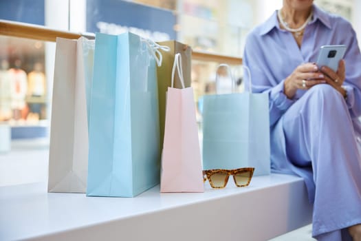 Mature woman resting after shopping sitting on bench in supermarket and chatting social media with friends or calling taxi cab using smartphone. Crop view