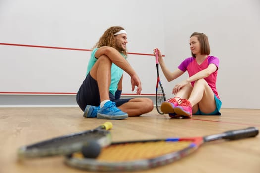 Male and female squash players taking break sitting on floor while rest and relax. Handsome sportive people and successful recreational pursuit at sport club