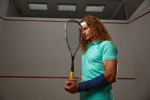 Young pensive racketball player with racket standing backside. Squash game practice at indoor court. Professional sport and hobby concept