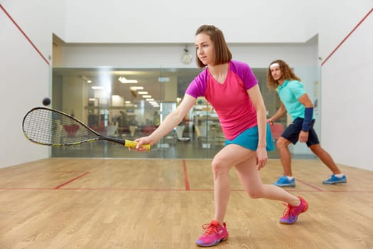 Young sportive couple practicing squash at sport club indoor court. Focus on attractive woman hitting ball with racket