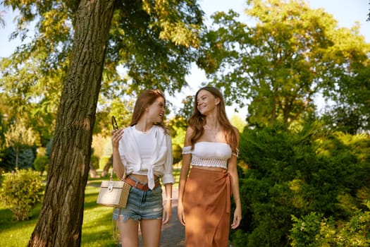 Two beautiful young girl friends wearing trendy fashion outfit walking in green summer public park. Attractive female teenagers laughing talking and sharing positive emotions