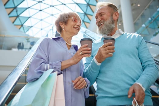 Senior couple with purchases in bags and coffee caps riding down shopping mall escalator enjoying time together