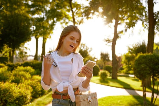 Portrait of young pretty woman with smartphone in park at sunny day. Happy cheerful teenager girl with look fashion