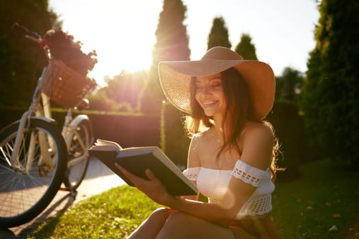 Charming young woman reading book while sitting on park bench in sunlight beam on sunset. Cheerful teenage female relaxing enjoying happy end of beautiful day outdoors