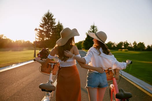 View from back on two female friends exploring nature on bikes outdoors. Hipster teenage women walking and hugging together sharing support and trust