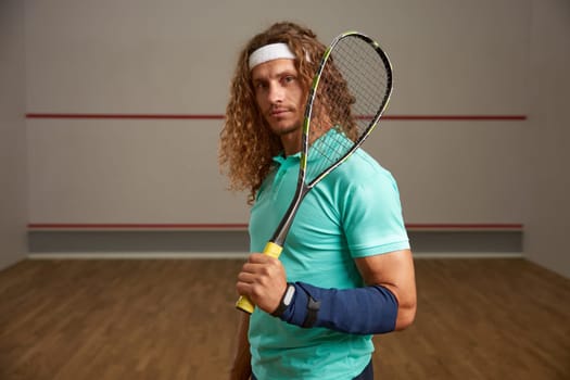 Portrait of confident male squash player standing with racket ready for game looking confidently at camera