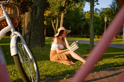 Charming young woman reading book while sitting in park on lawn in sunlight beam on sunset. Cheerful teenage female relaxing enjoying happy end of beautiful day outdoors