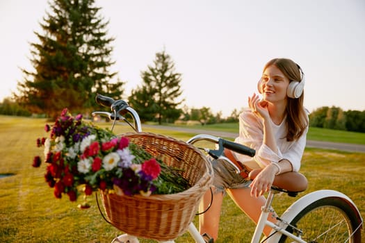 Young hipster woman florist wearing headphones listening to music resting after long bicycle race for wildflowers on green field. Craft hobby and traveling outdoors