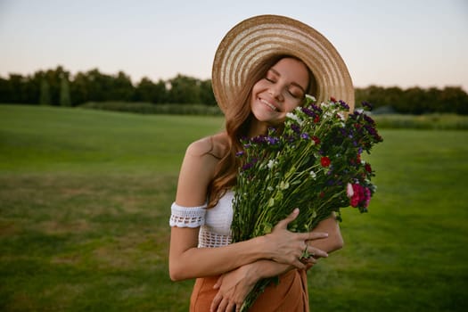 Beautiful woman holding bunch of wildflower standing over green countryside field. Femininity, natural beauty and innocence concept