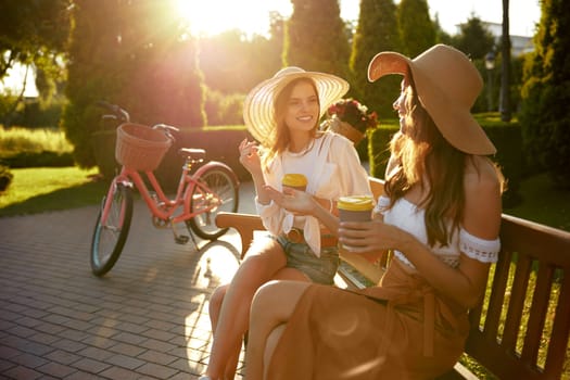 Two gorgeous women friends in sunlight beams sitting on park bench chatting while drinking takeaway coffee after active bicycle riding