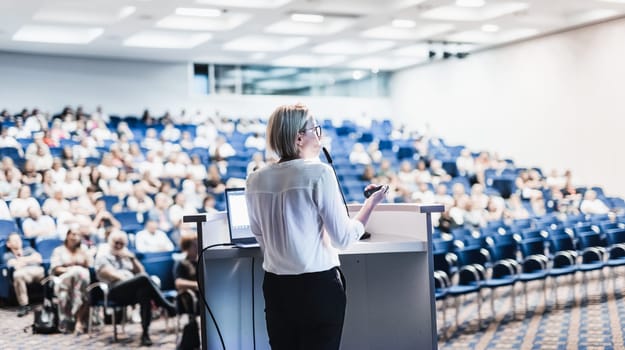Female speaker giving a talk on corporate business conference. Unrecognizable people in audience at conference hall. Business and Entrepreneurship event