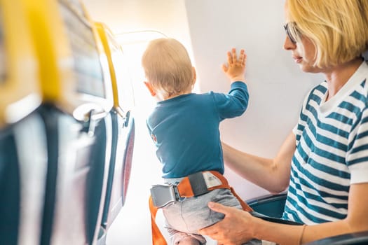 Mom and child flying by plane. Mother holding and playing with her infant baby boy child in her lap during economy comercial flight. Concept photo of air travel with baby. Real people