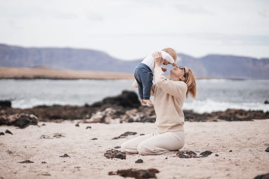 Mother enjoying winter vacations holding, playing and lifting his infant baby boy son high in the air on sandy beach on Lanzarote island, Spain. Family travel and vacations concept.