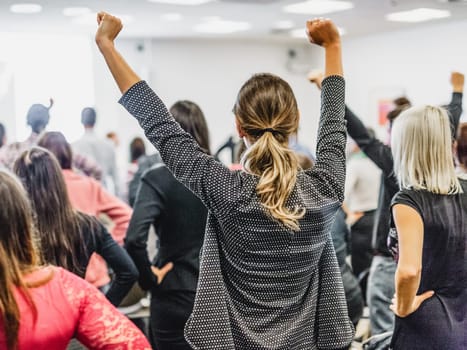 Life coaching symposium. Speaker giving interactive motivational speech at business workshop. Rear view of unrecognizable participants feeling empowered and motivated, hands raised high in air.