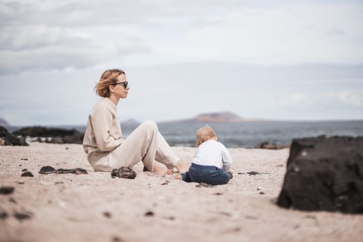 Mother enjoying winter beach vacations playing with his infant baby boy son on wild volcanic sandy beach on Lanzarote island, Canary Islands, Spain. Family travel and vacations concept.