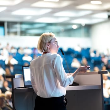 Female speaker giving a talk on corporate business conference. Unrecognizable people in audience at conference hall. Business and Entrepreneurship event