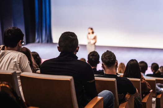 Business and entrepreneurship symposium. Female speaker giving a talk at business meeting. Audience in conference hall. Rear view of unrecognized participant in audience.