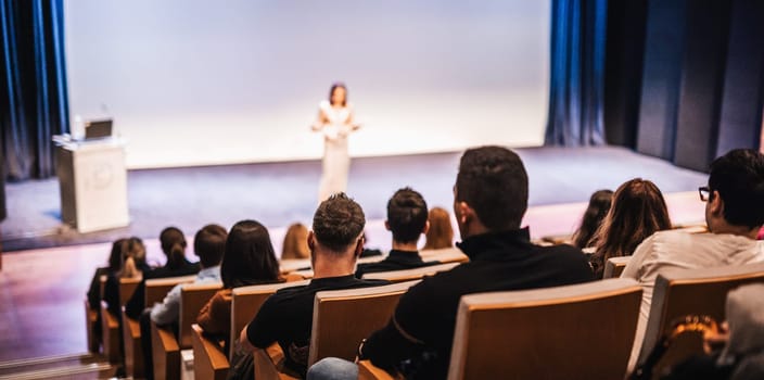 Business and entrepreneurship symposium. Female speaker giving a talk at business meeting. Audience in conference hall. Rear view of unrecognized participant in audience.