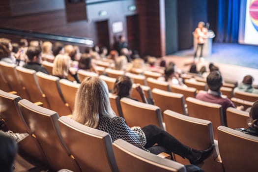 Business and entrepreneurship symposium. Female speaker giving a talk at business meeting. Audience in conference hall. Rear view of unrecognized participant in audience.