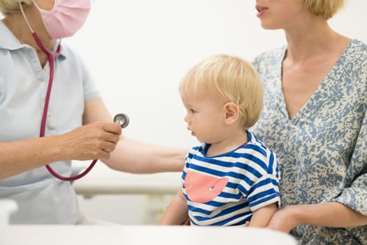 Infant baby boy child being examined by his pediatrician doctor during a standard medical checkup in presence and comfort of his mother. National public health and childs care care koncept