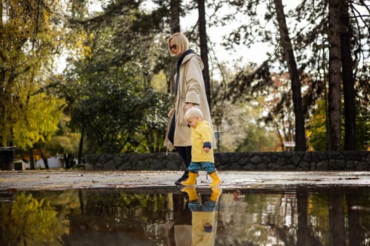 Small bond infant boy wearing yellow rubber boots and yellow waterproof raincoat walking in puddles on a overcast rainy day holding her mother's hand. Mom with small child in rain outdoors
