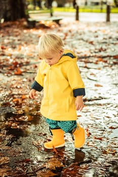 Small blond infant boy wearing yellow rubber boots and yellow waterproof raincoat walking in puddles on a overcast rainy day. Child in the rain