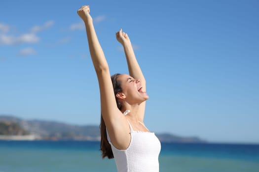 Excited woman in white dress celebrating raising arms on the beach a sunny summer day