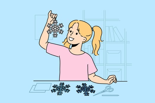 Little girl cuts out paper snowflakes preparing handmade christmas decorations for home on eve of new year. Child stands near table with scissors demonstrating snowflake made with own hands