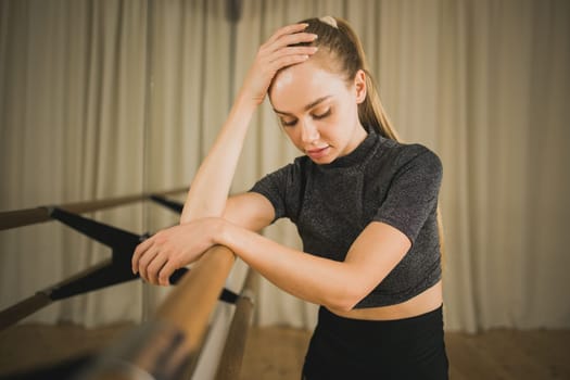 Upset ballerina in training suit stands near ballet barre and looks at the mirror in dance studio. Woman prepares for performance. Ballet dancer doing gymnastic exercises. Classical ballet school.