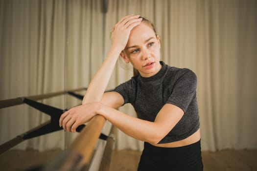 Upset ballerina in training suit stands near ballet barre and looks at the mirror in dance studio. Woman prepares for performance. Ballet dancer doing gymnastic exercises. Classical ballet school.