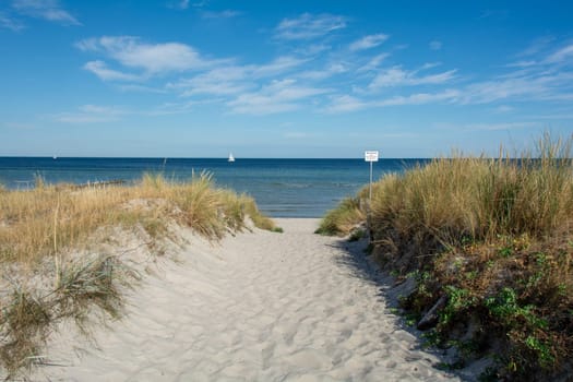 Path between the sand dunes overlooking the sea and a sailing ship and blue sky. With sign "Grones - Please keep your distance - risk of accident"""