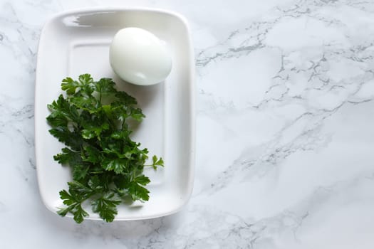 Boiled chicken egg and parsley on a white plate on a marble background.