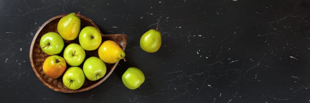 Wooden carved bowl with pears and apples on black marble board photo from above. Wide banner space for text right