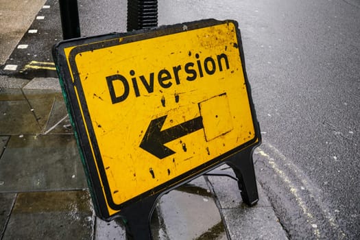 Yellow diversion sign with arrow pointing left on wet asphalt road.