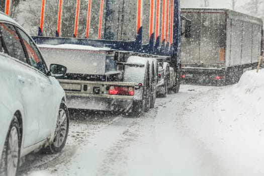 Car waiting behind large trucks during traffic jam on forest road, caused by heavy snow blizzard