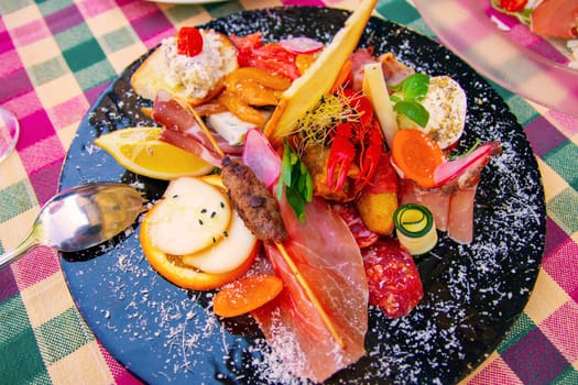 Traditional Italian food. Close-up of mixed Mediterranean appetizers or antipasto and tapas with ham vegetables and crab on black plate on restaurant table.