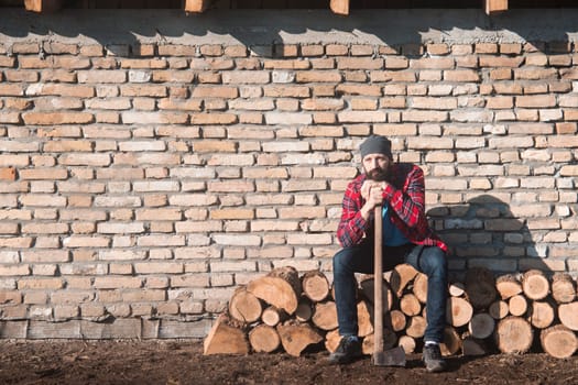 The sad and tired lumberjack worker with axe is sitting on firewood on brick wall.