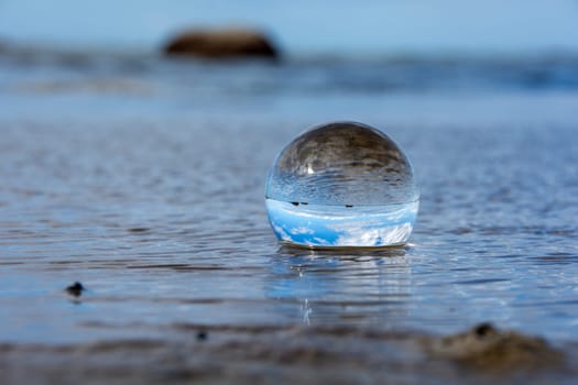A glass ball lies in the waves on the sandy beach, the sea and the setting sun are reflected in the ball