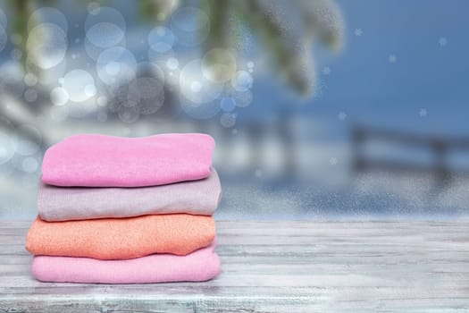 Pile of colorful knitted cozy warm cashmere sweaters on wooden table against blurred abstract snowy landscape xmas background. Template for christmas sale.