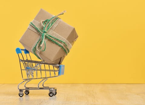 A gift wrapped in brown craft paper in a miniature shopping cart on a yellow background