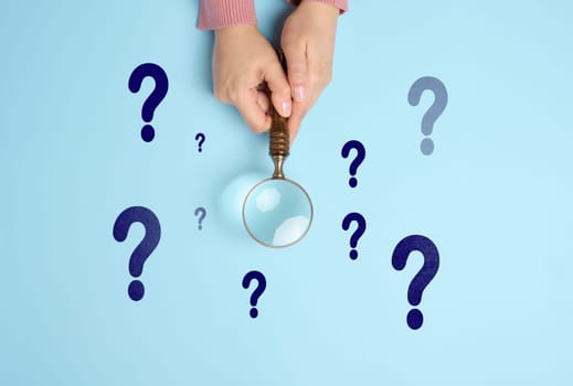 Female hand holds a wooden magnifying glass and question marks on a blue background. The concept of finding an answer to questions, truth and uncertainty.