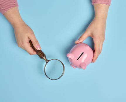 A woman's hand holds a piggy bank and a magnifying glass on a blue background, top view