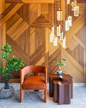 A simple and elegant image of a modern design orange chair and coffe table in front of a decorated wood cladding wall, with hanging lights and a potted plant
