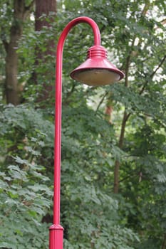 a red street lamp stands in front of a vegetated background