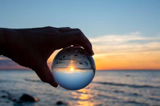 A hand of an elderly person holds a glass ball, the sea and the setting sun are reflected in the ball