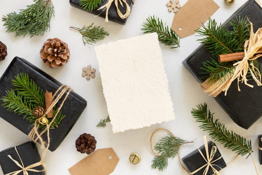 Paper card between Christmas decor, black gift boxes and green fir branches top view. Vertical blank card mockup, flat lay, copy space. Christmas or New Year Holiday greeting