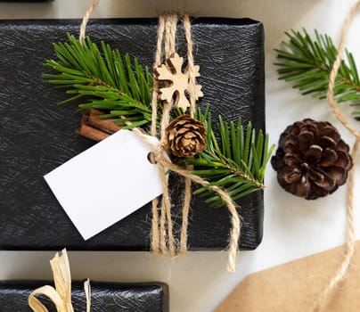 Black wrapped present with Christmas gift tag with fir tree branches, pine cones and holiday decorations, top view. Rustic winter composition with blank Gift tag Mockup, copy space, close up