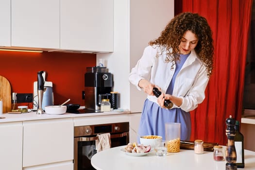 Young housewife adding pepper into blender with chickpeas while making hummus