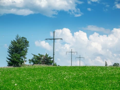 Overhead power line with four masts on a hill in the Allgäu in summer in front of a blue sky with white clouds.