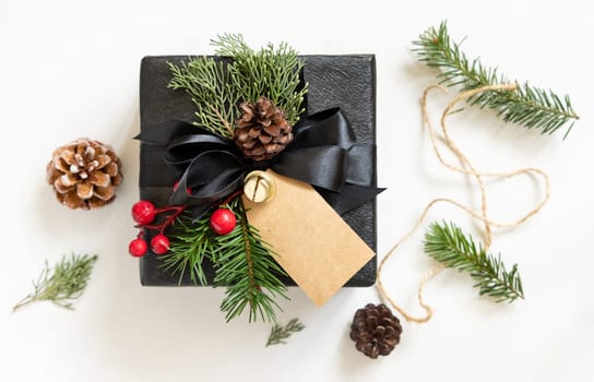 Black present with Christmas vertical gift tag, fir tree branches, pine cones and holiday decorations, top view. Winter composition with blank label Mockup, copy space, top view